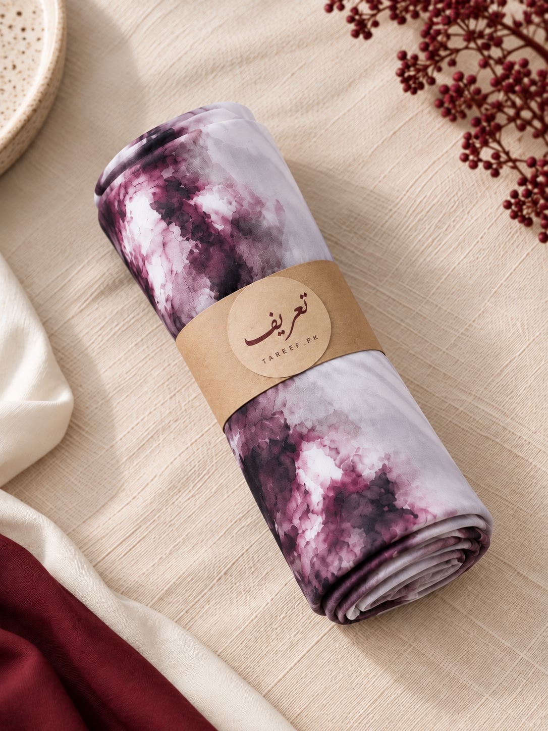 A lightweight hijab with a purple and white watercolor-like print, styled on a white mannequin wearing a white dress in a warm, sunlit interior setting.