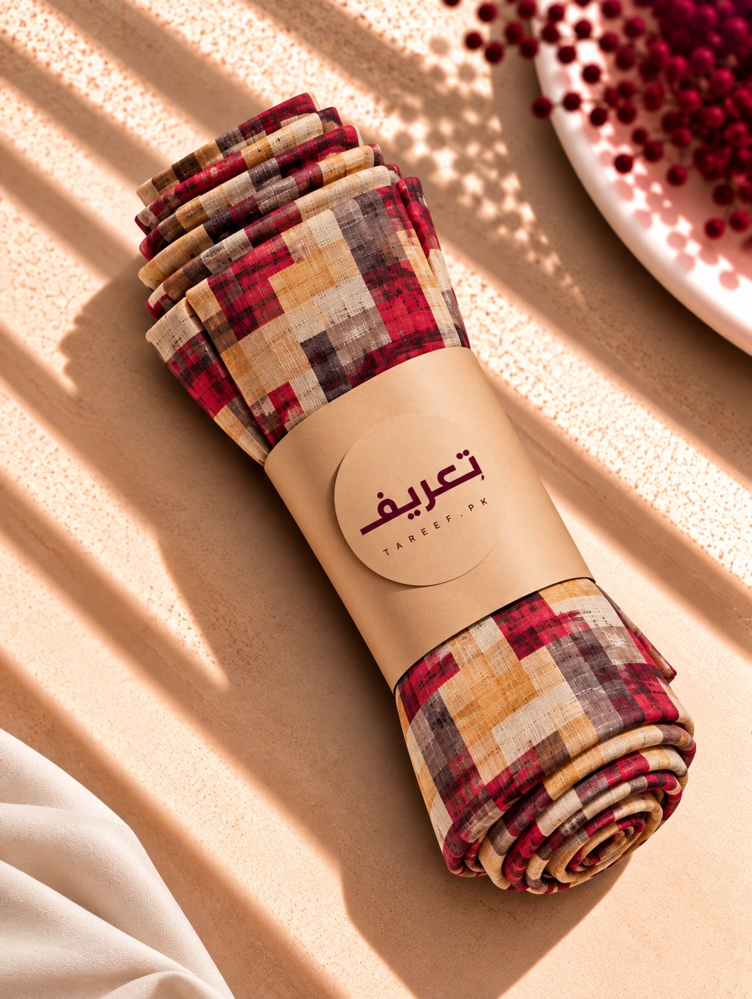 A rolled hijab wrapped in a kraft paper band with Arabic text reading 'TAREEf' placed on a textured beige surface with warm sunlight shadows.