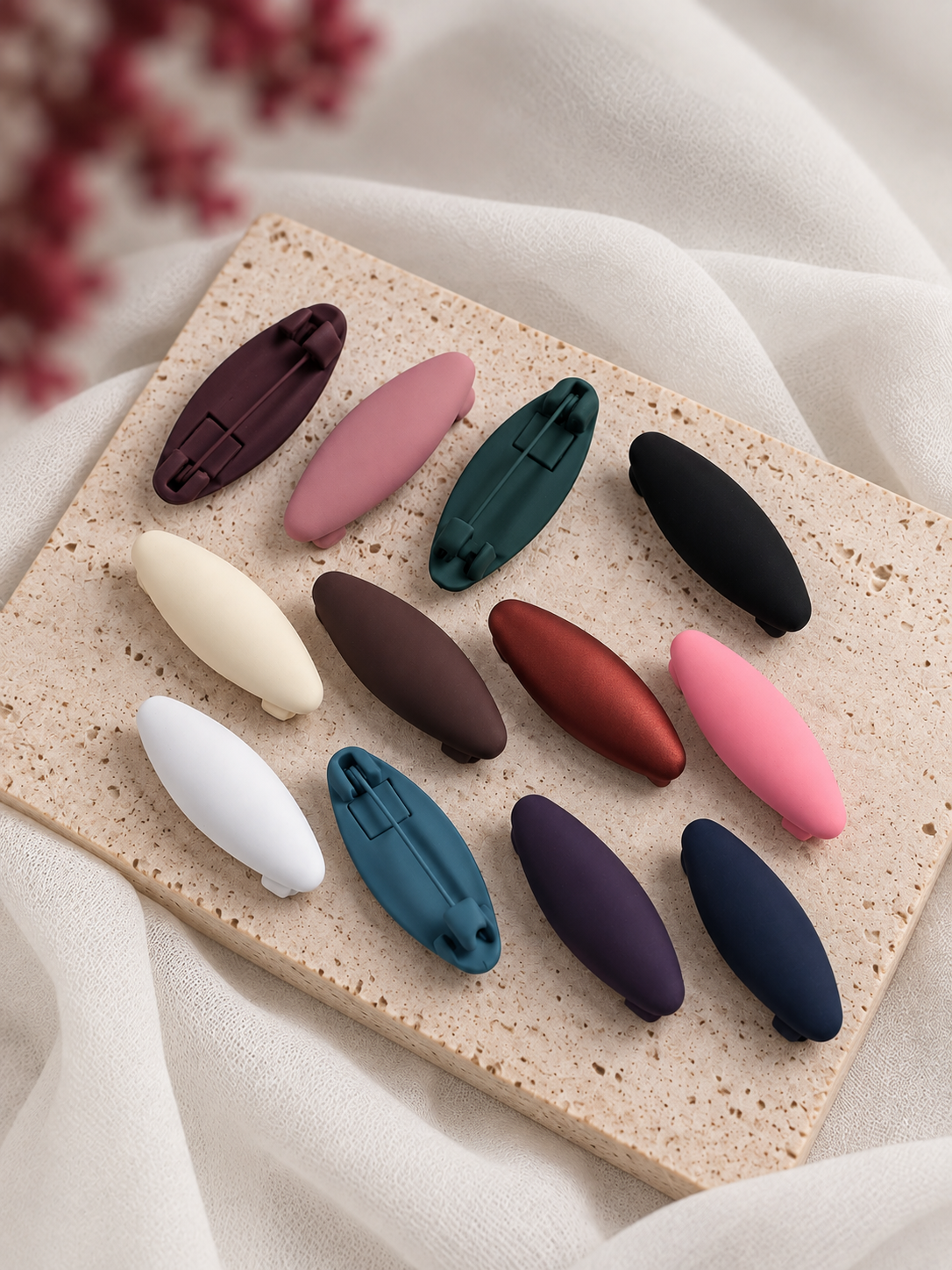 Four oval hair matte hijab clips in colors (pink, purple, navy, and brown) arranged on a textured beige stone slab with draped white fabric and dried flowers.