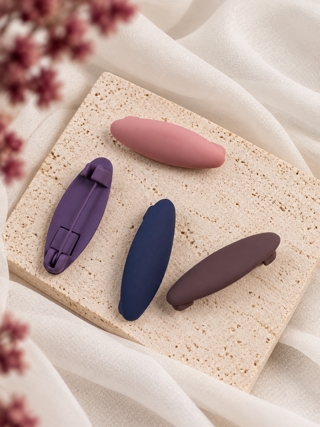 Four oval hair matte hijab clips in colors (pink, purple, navy, and brown) arranged on a textured beige stone slab with draped white fabric and dried flowers.