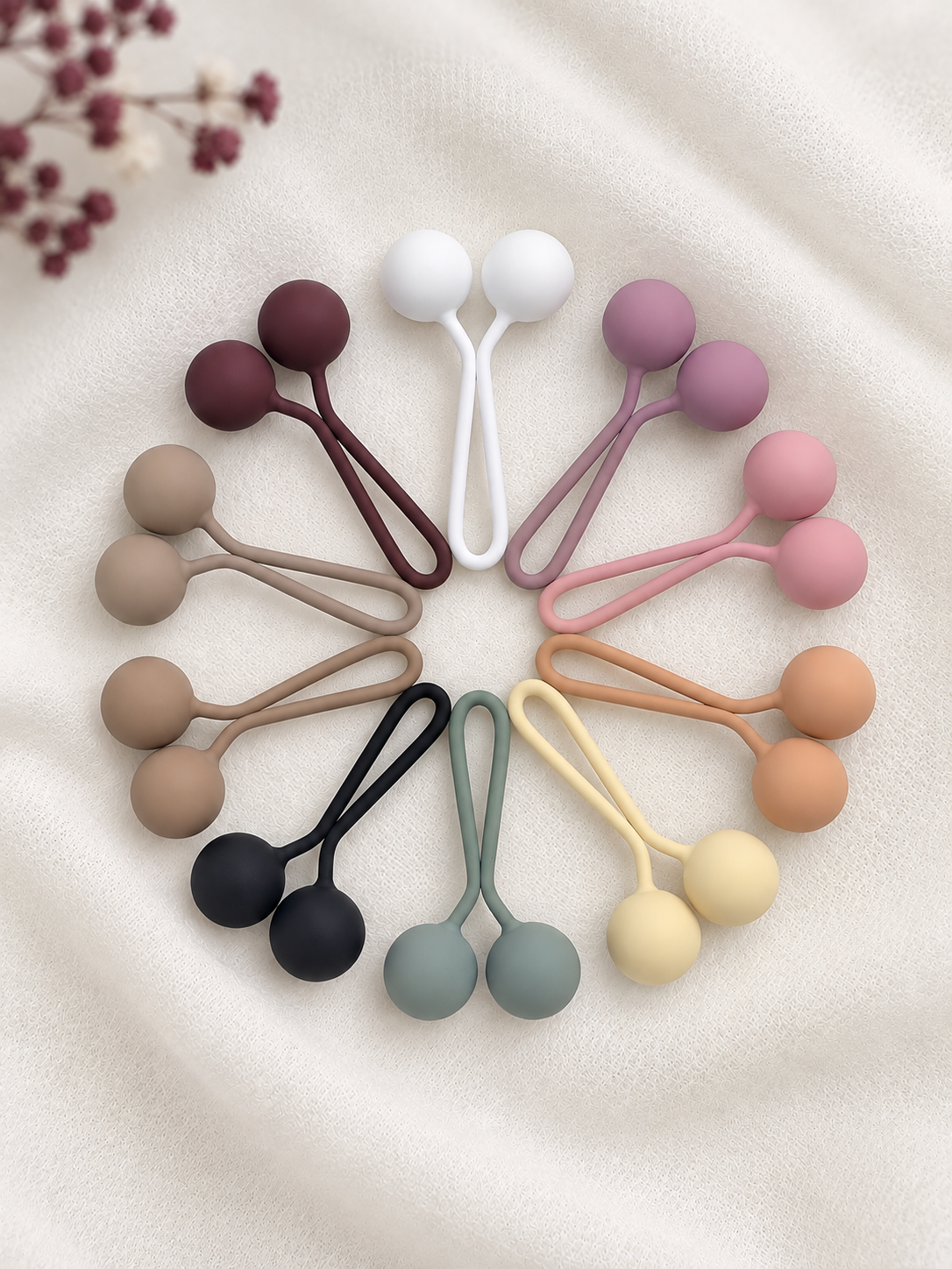 Ten matte hijab loop pins of silicone arranged in a circular flat lay on a soft cream fabric background. The set includes white, burgundy, mauve, pink, beige, black, sage green, yellow, and peach tones.