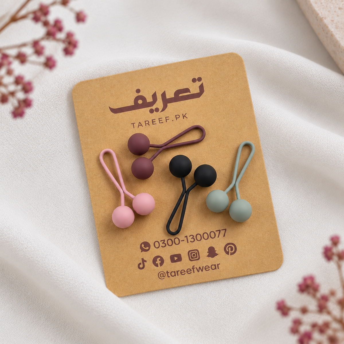 Four matte hijab loop pins in pink, black, gray, and maroon are displayed on a brown kraft card with the brand name 'TAREEF' styled on a white fabric background with dried flowers.