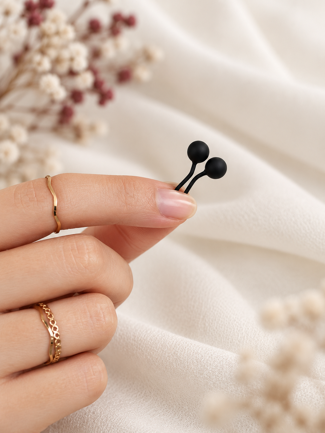 A close-up of a hand holding a matte hijab loop pins against a soft cream fabric backdrop with dried flowers.