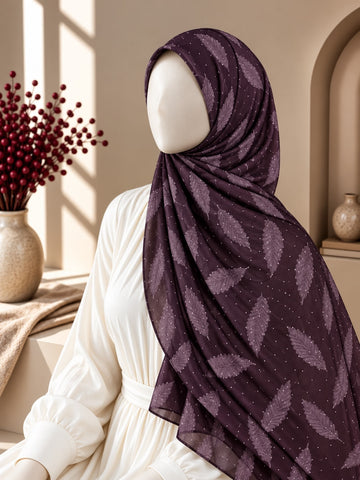A draped hijab in deep plum purple with a tonal leaf pattern, styled on a mannequin wearing a white dress in a warm, sunlit interior setting.