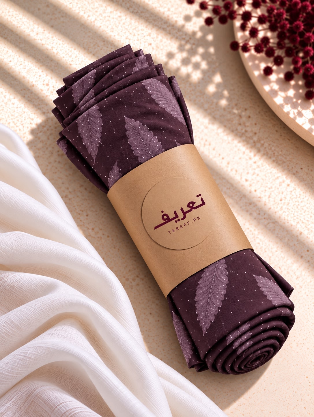 A rolled dark purple hijab with a leaf pattern and tiny white dots, wrapped in a brown paper band with the brand name 'TAREEF' styled on a textured beige surface with draped white fabric and shadows.