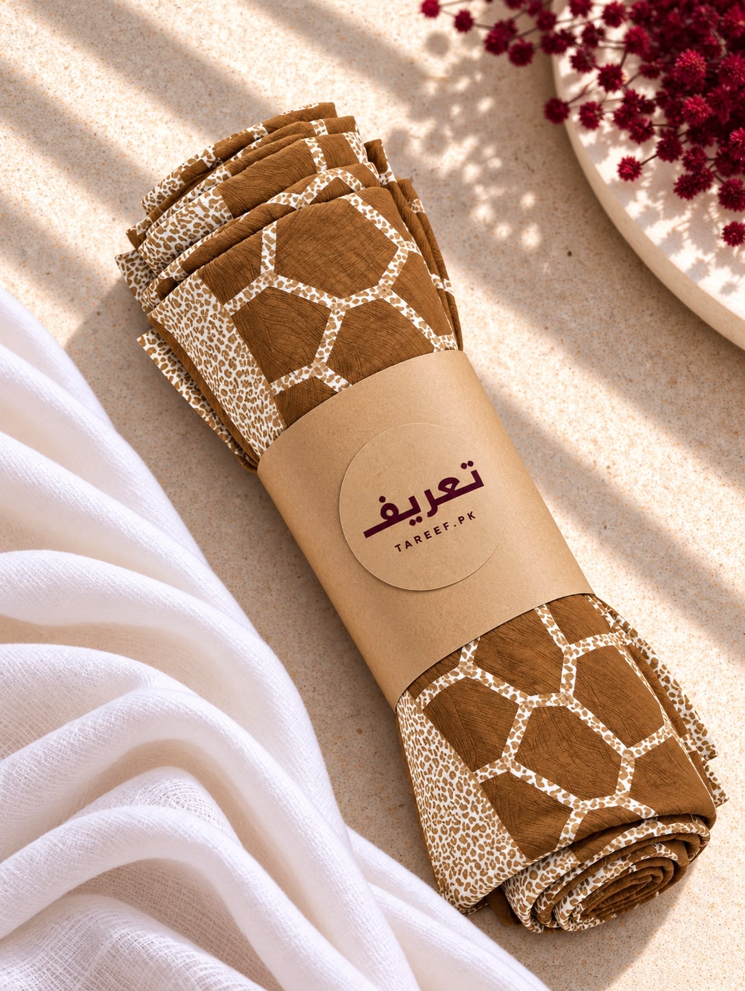 A rolled hijab with a brown and beige giraffe-like pattern, wrapped in a kraft paper band labeled 'TAREEF,' styled on a light beige surface with draped white fabric and red flowers in the corner.