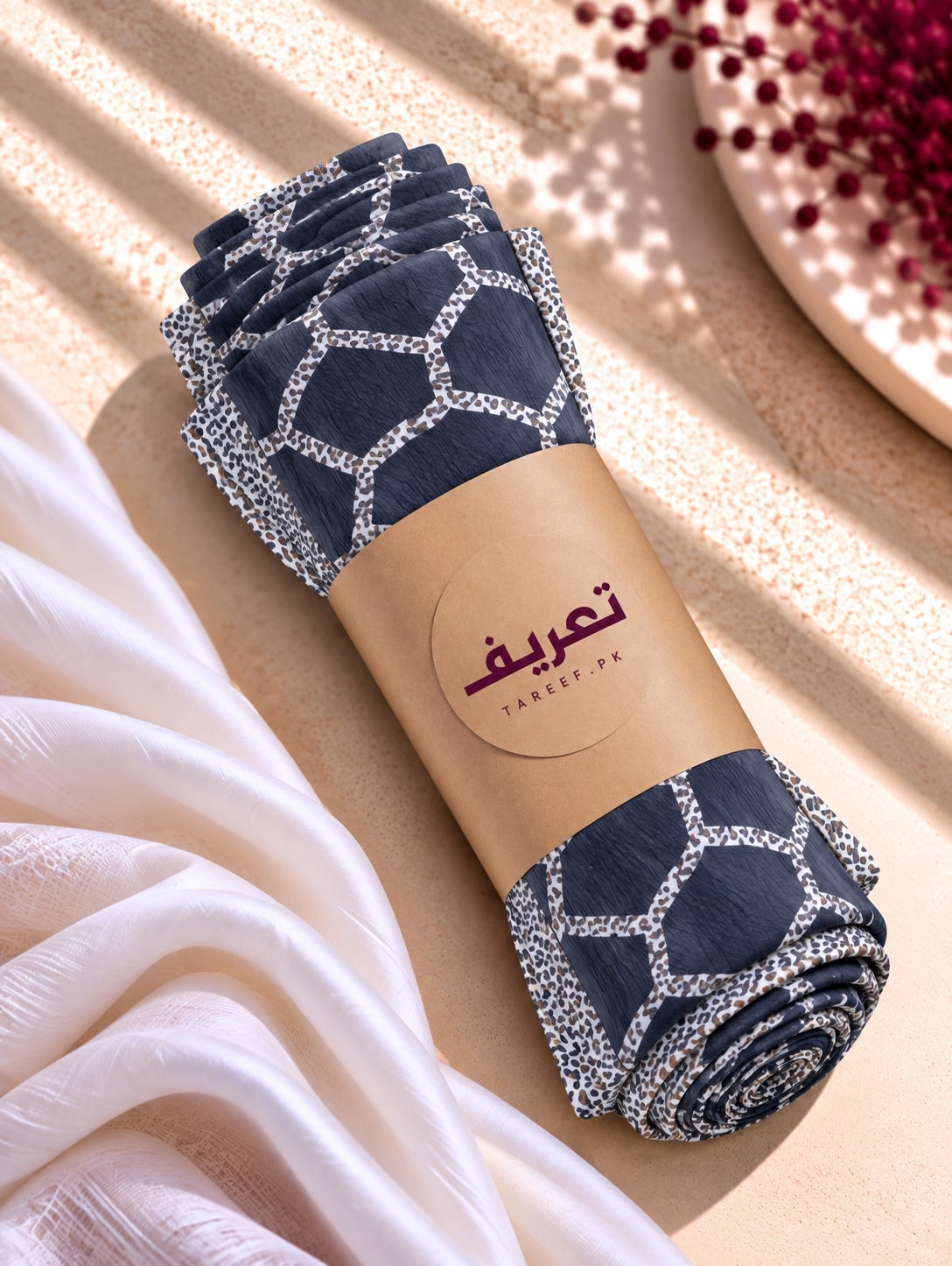 A navy blue hijab with a white geometric lattice pattern and a matching patterned border, styled on a mannequin wearing a white long-sleeve dress in a warm, sunlit interior setting.
