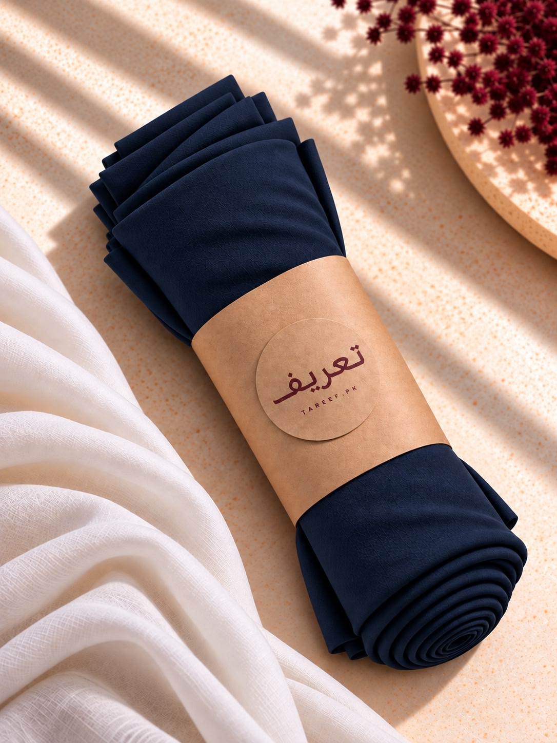 A rolled navy blue hijab wrapped in a kraft paper band with a circular label 'TAREEF', styled on a warm beige surface with draped white fabric and dried red flowers.
