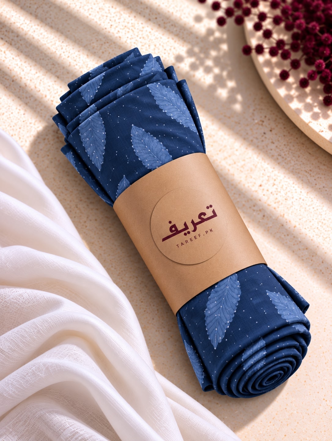 A rolled navy blue hijab with a light blue leaf pattern and tiny white dots, wrapped in a tan paper band printed with the brand name 'TAREEF' styled on a textured beige surface with draped white fabric and decorative red flowers.