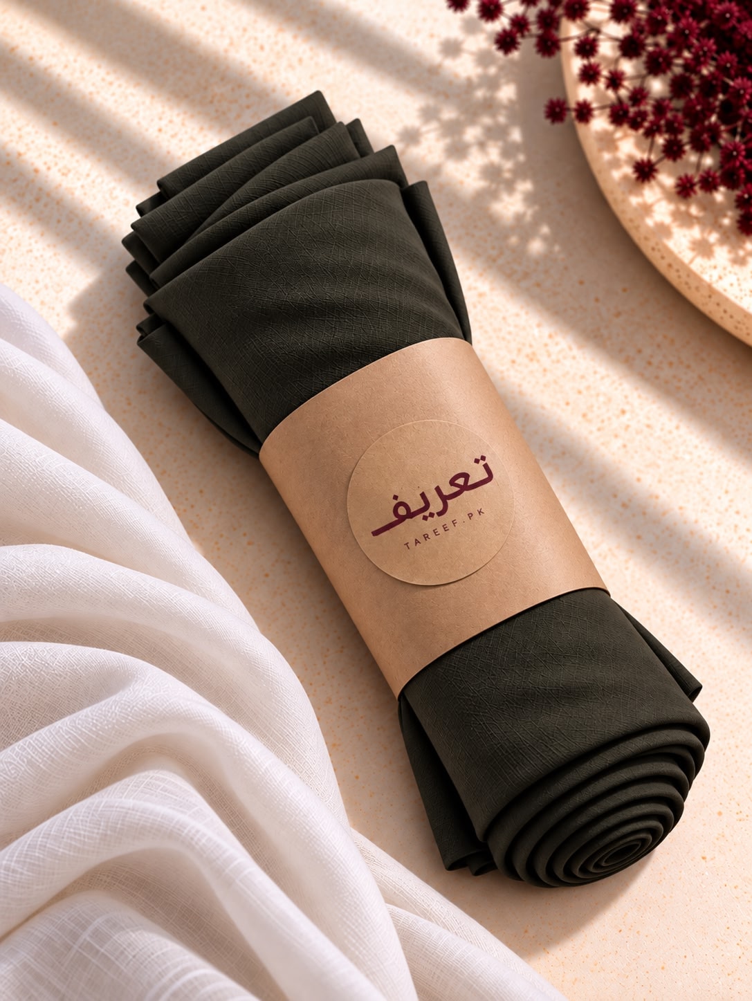 A rolled dark olive green hijab wrapped in a kraft paper band 'TAREEF' printed on it, styled on a beige textured surface with draped white fabric and red flowers.
