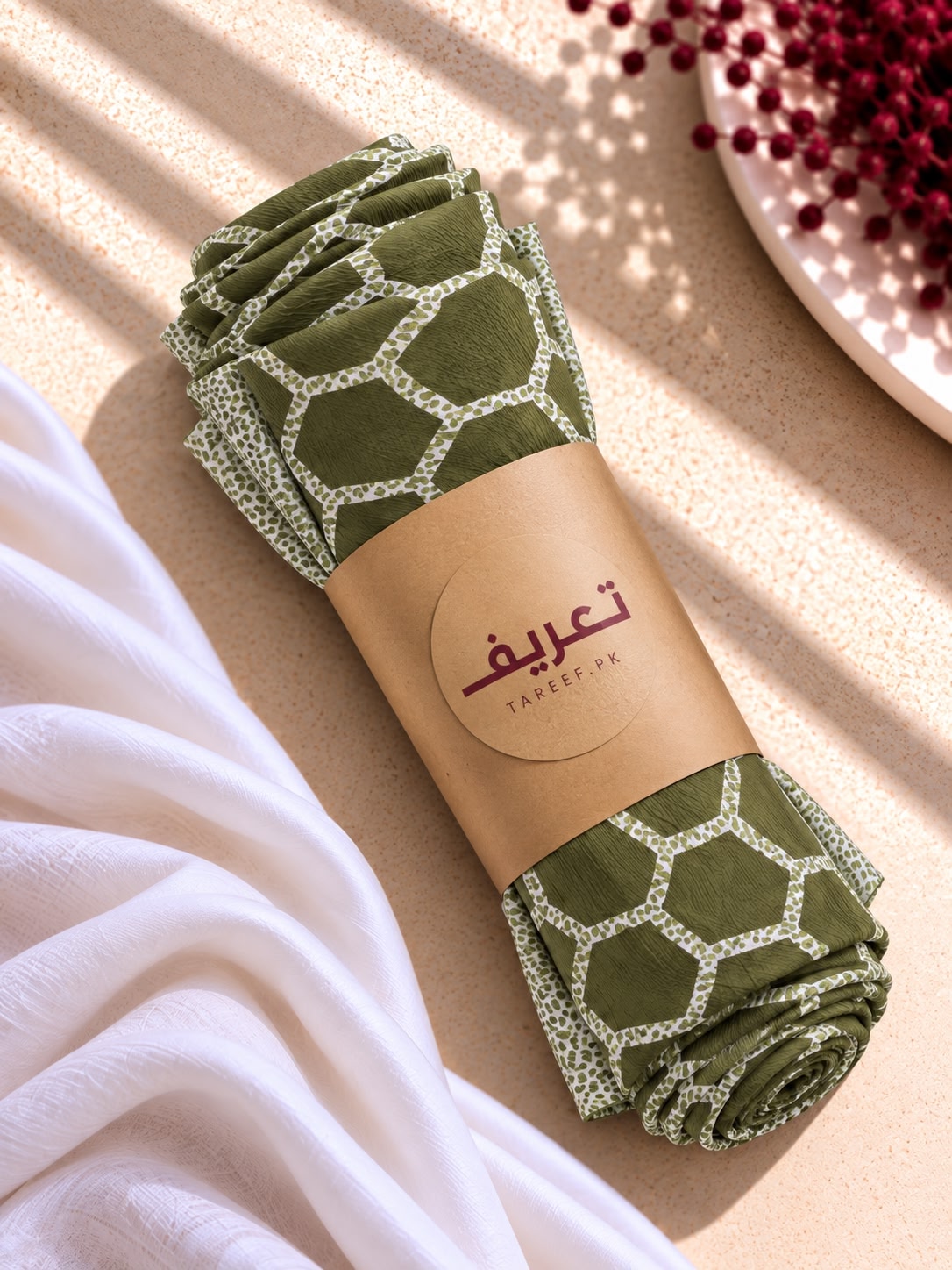 A rolled olive green hijab with a white geometric pattern, wrapped in a brown paper band with the brand name 'TAREEF' styled on a beige textured surface with white draped fabric and red berries.
