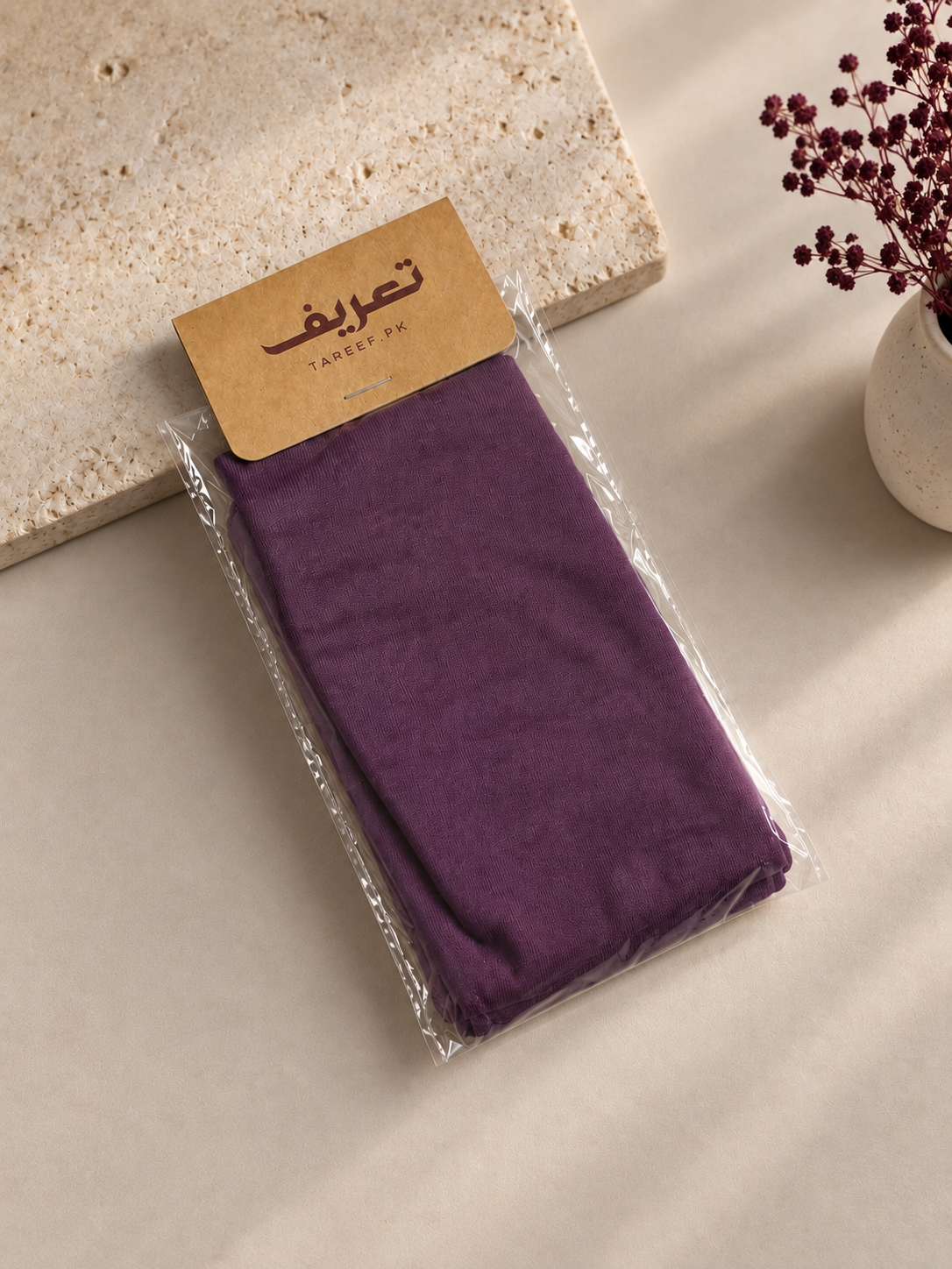 A folded purple hijab cap in clear plastic packaging with a tan paper label that reads 'TAREEF' styled on a beige surface with a stone slab and dried flowers in a vase.