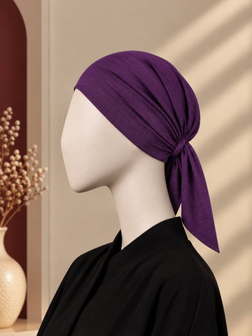 A purple hijab cap tied in a knot, displayed on a white mannequin head wearing a black top, set against a warm beige background with soft window shadow patterns.
