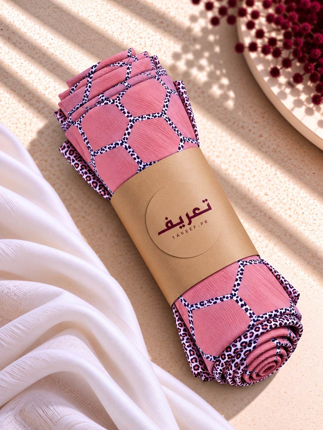 A rolled pink hijab with a geometric and leopard print pattern, wrapped in a brown paper band 'TAREEF,' styled on a textured beige surface with draped white fabric and sunlit shadows.