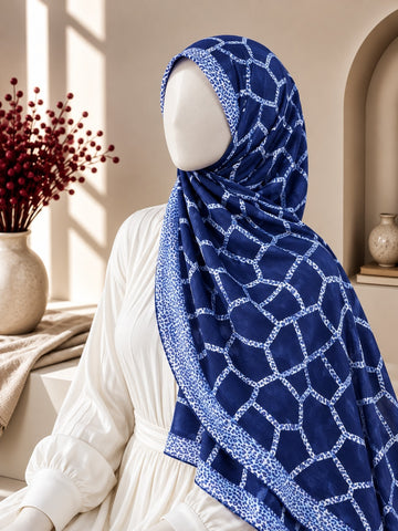 A navy blue hijab with a white geometric lattice pattern and matching border, styled on a mannequin wearing a white dress in a warm, sunlit interior setting.