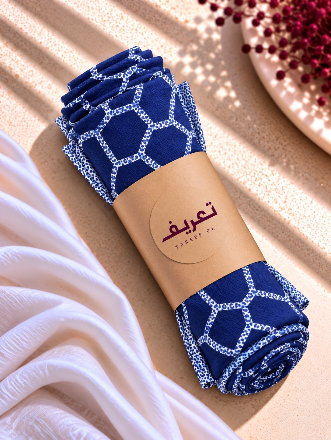A navy blue hijab with a white geometric lattice pattern and matching border, styled on a mannequin wearing a white dress in a warm, sunlit interior setting.