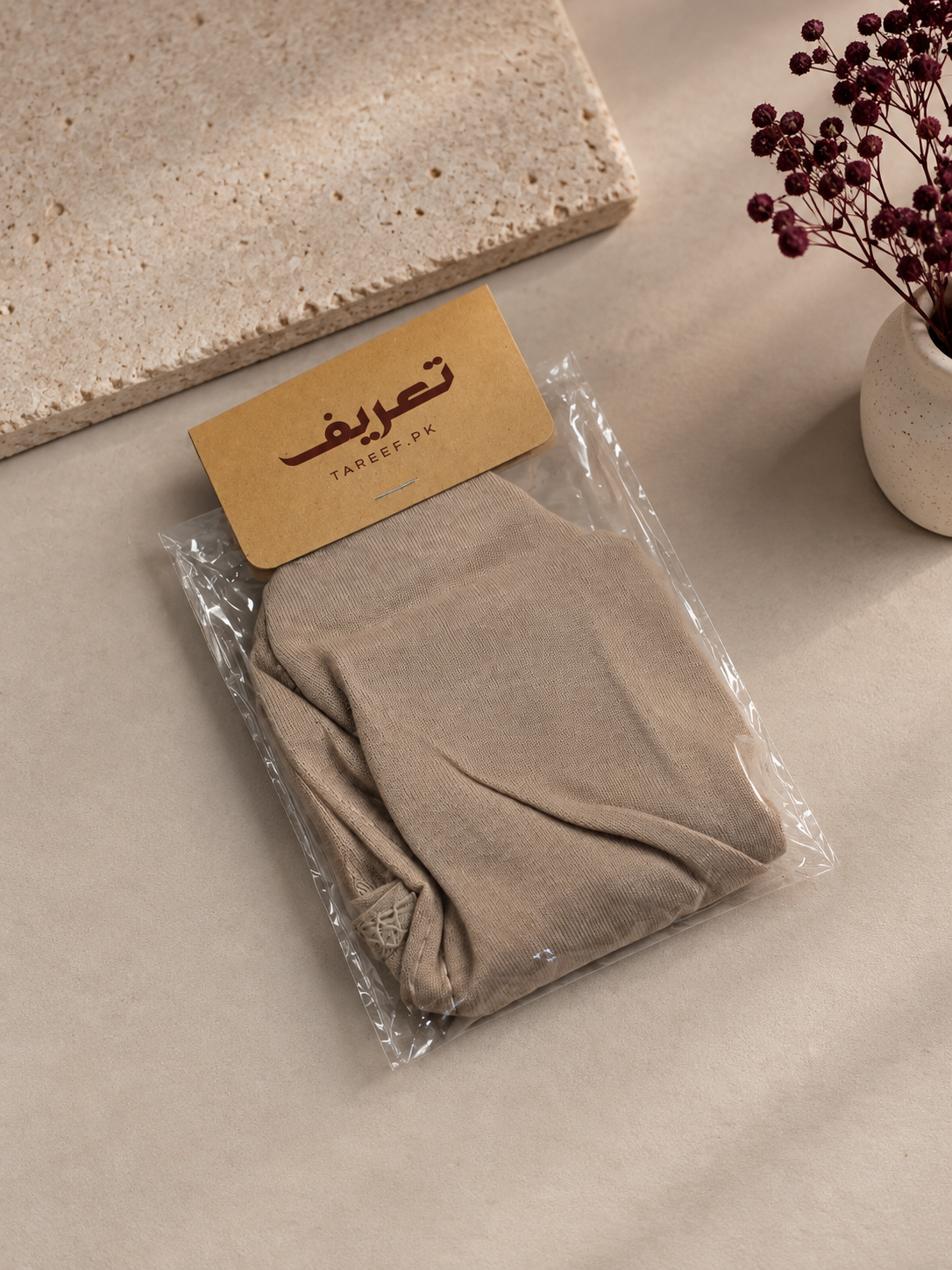 A light taupe knotted hijab cap displayed on a white mannequin head, styled with a black top against a warm beige background with soft window shadow patterns.