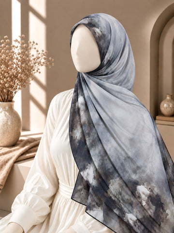 A mannequin wearing a large light blue-gray hijab with a dark navy watercolor-style print, posed in a warm beige interior with dried flowers and soft window light.