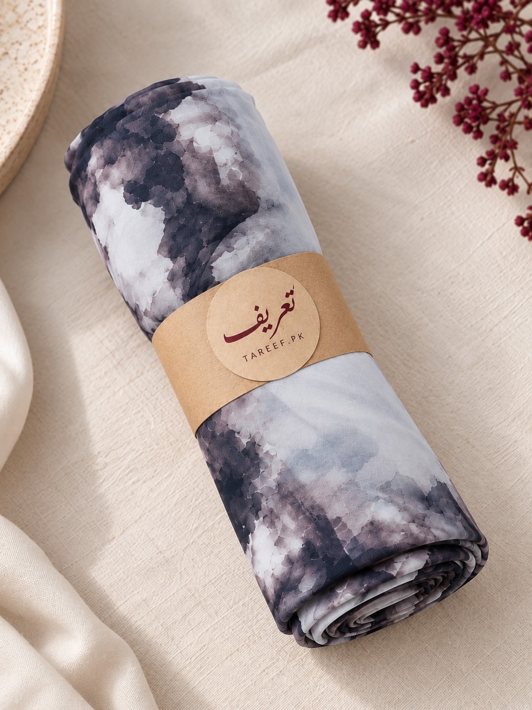 A rolled hijab with a gray, white, and black watercolor-style print, wrapped in a tan paper band labeled 'TAREEF,' styled on a beige fabric surface with dried flowers and a woven mat edge.