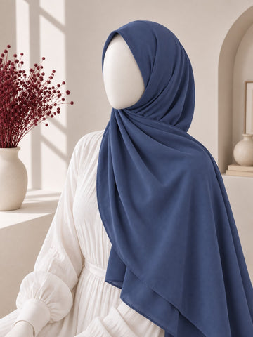 A navy blue hijab draped elegantly on a white mannequin wearing a white dress, set in a bright, modern interior with warm sunlight and a vase of red dried flowers.
