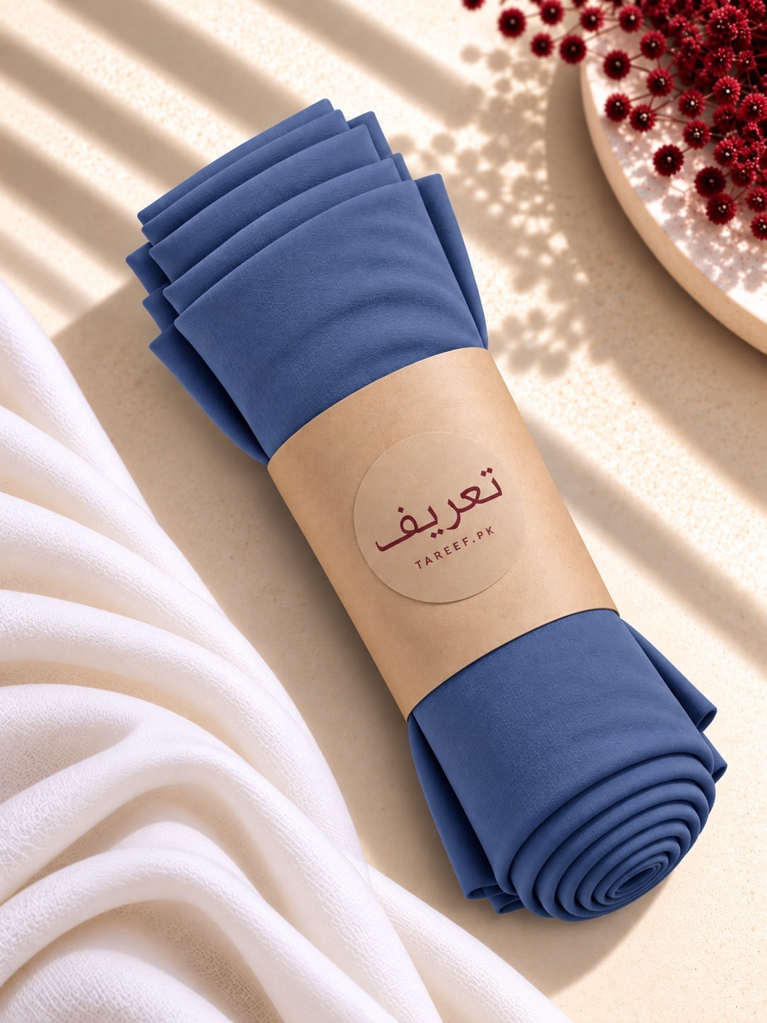 A rolled bundle of blue hijab with a beige paper band labeled 'TAREEF' styled on a light beige surface with draped white fabric and red decorative beads in the corner.