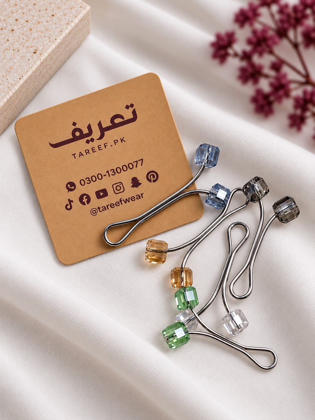 Ten curved hijab loop pins with translucent colored beads in various colors, arranged in a circular pattern on a soft white fabric background.