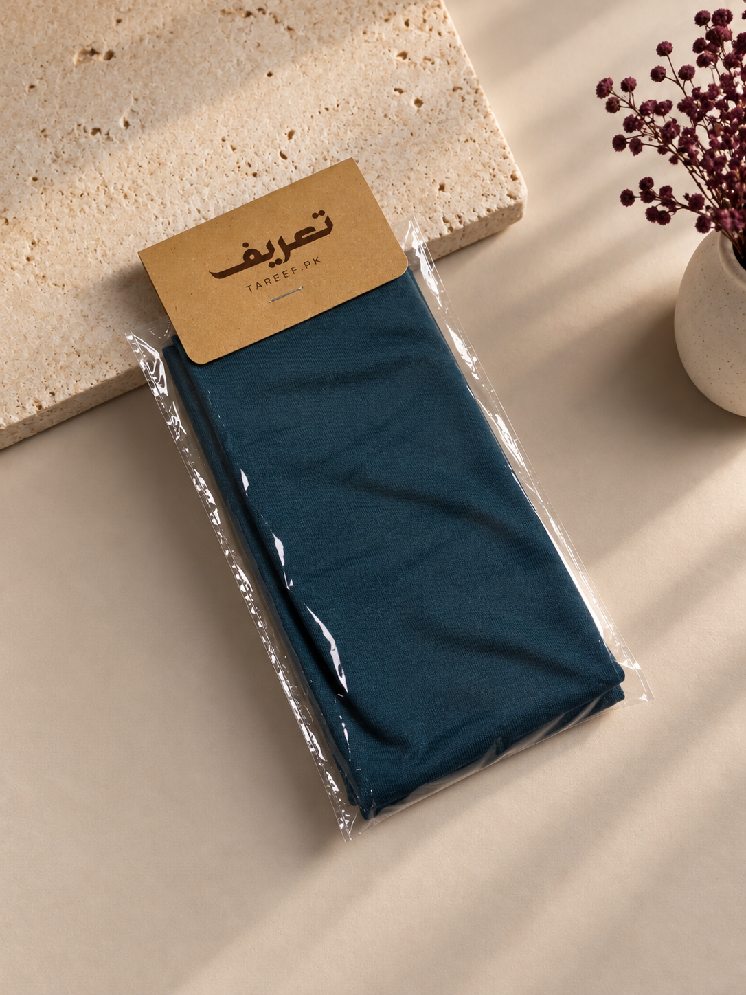 A teal blue hijab cap tied in a knot on a white mannequin head, styled with a black top against a warm beige background with soft window shadow patterns.