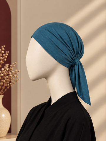 A teal blue hijab cap tied in a knot on a white mannequin head, styled with a black top against a warm beige background with soft window shadow patterns.