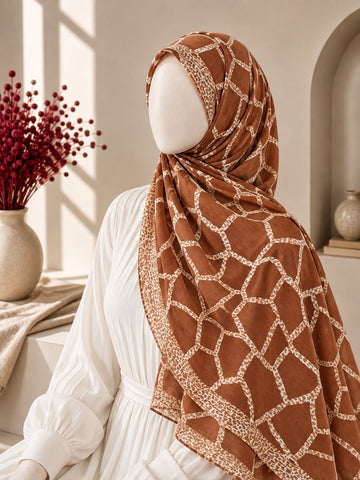 A terracotta brown hijab with a beige geometric lattice pattern and matching border, styled on a white mannequin wearing a white dress in a warm, sunlit interior setting.