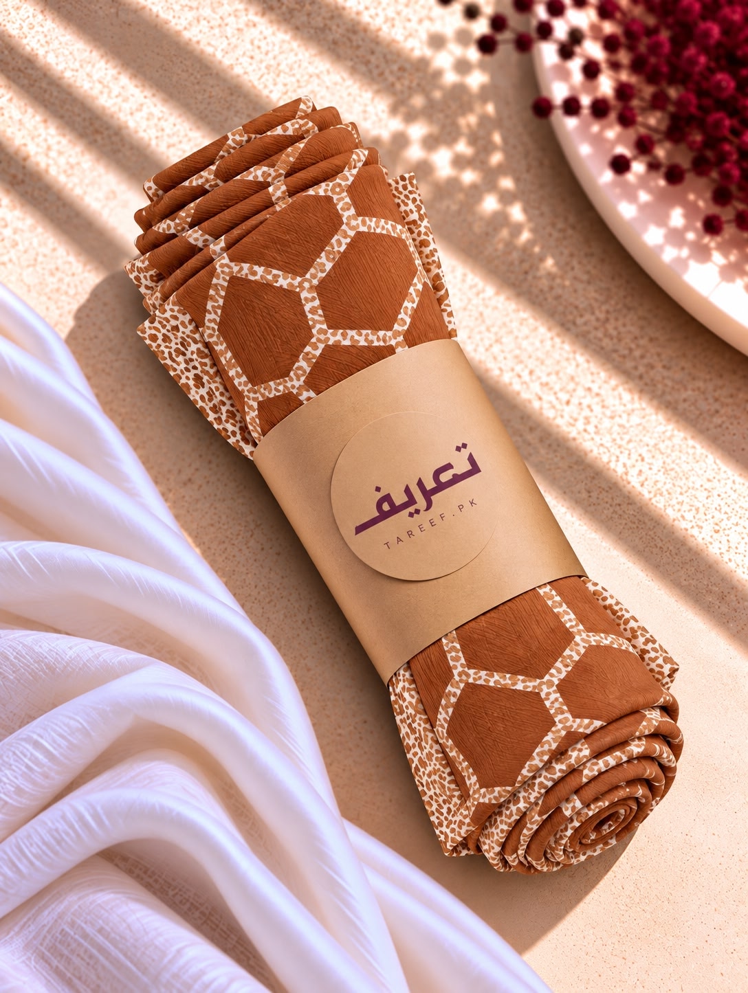 A rolled brown hijab with a beige geometric honeycomb pattern and a tan paper band labeled 'TAREEF' styled on a textured beige surface with draped white fabric and decorative red berries.