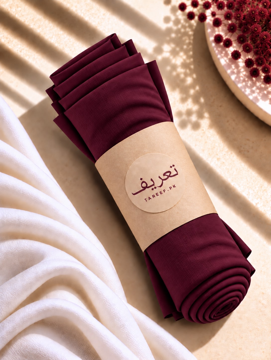 A deep burgundy hijab draped elegantly on a white mannequin wearing a white long-sleeve dress, set in a warm, softly lit interior with decorative vases and neutral walls.