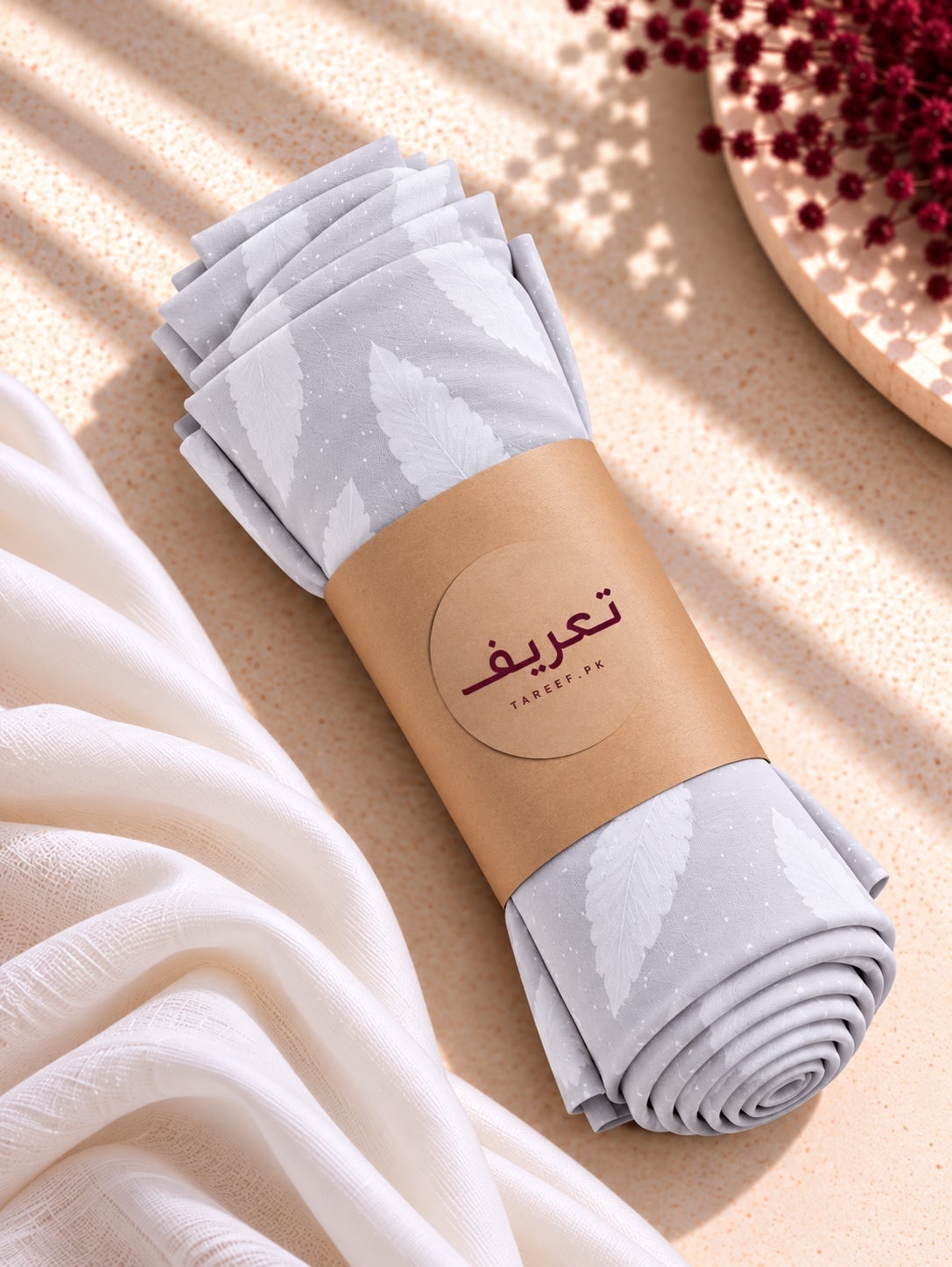 A rolled light gray hijab with a white leaf pattern, wrapped in a brown paper band labeled 'TAREEF,' styled on a beige textured surface with soft white fabric and dried red flowers.
