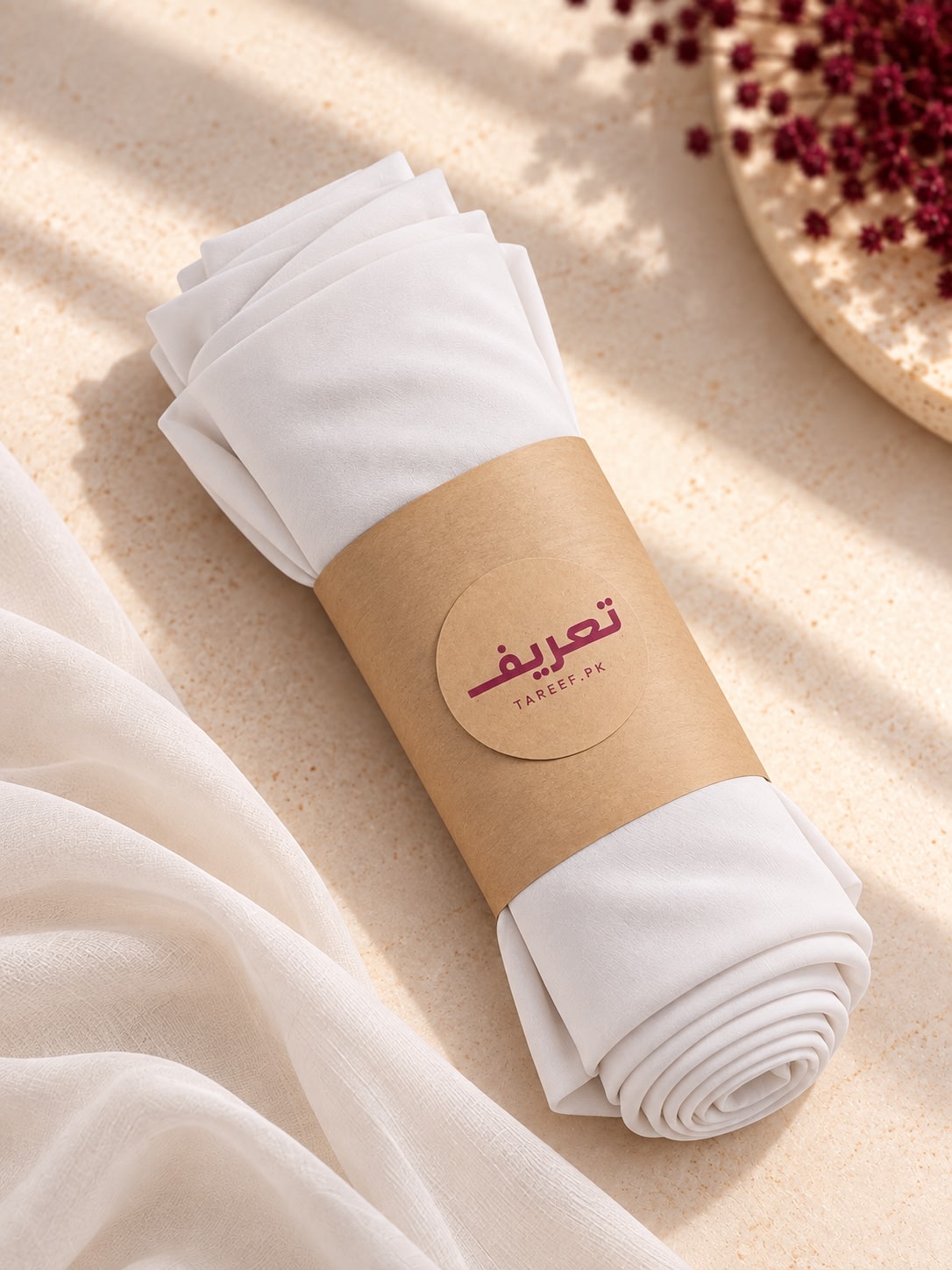 A white draped hijab and matching long-sleeve dress displayed on a mannequin in a warm, sunlit interior setting with beige walls, a window, and a vase of red berries.