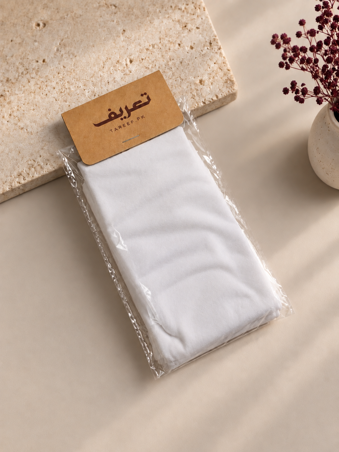 A folded white hijab cap in clear plastic packaging with a kraft paper label reading 'TAREEF' styled on a beige surface with a stone slab and dried flowers in the background.