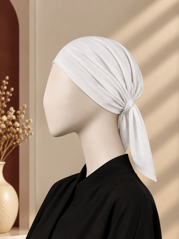A white hijab cap tied in a knot, displayed on a mannequin head wearing a black top, set against a warm beige background with soft window shadow patterns.