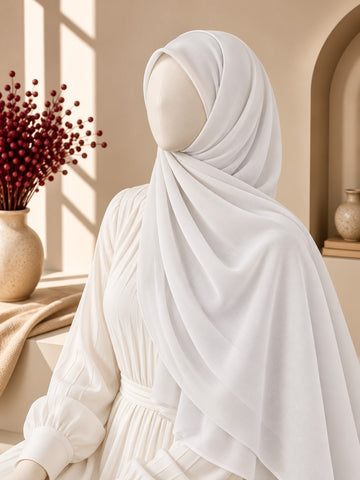 A white draped hijab and matching long-sleeve dress displayed on a mannequin in a warm, sunlit interior setting with beige walls, a window, and a vase of red berries.