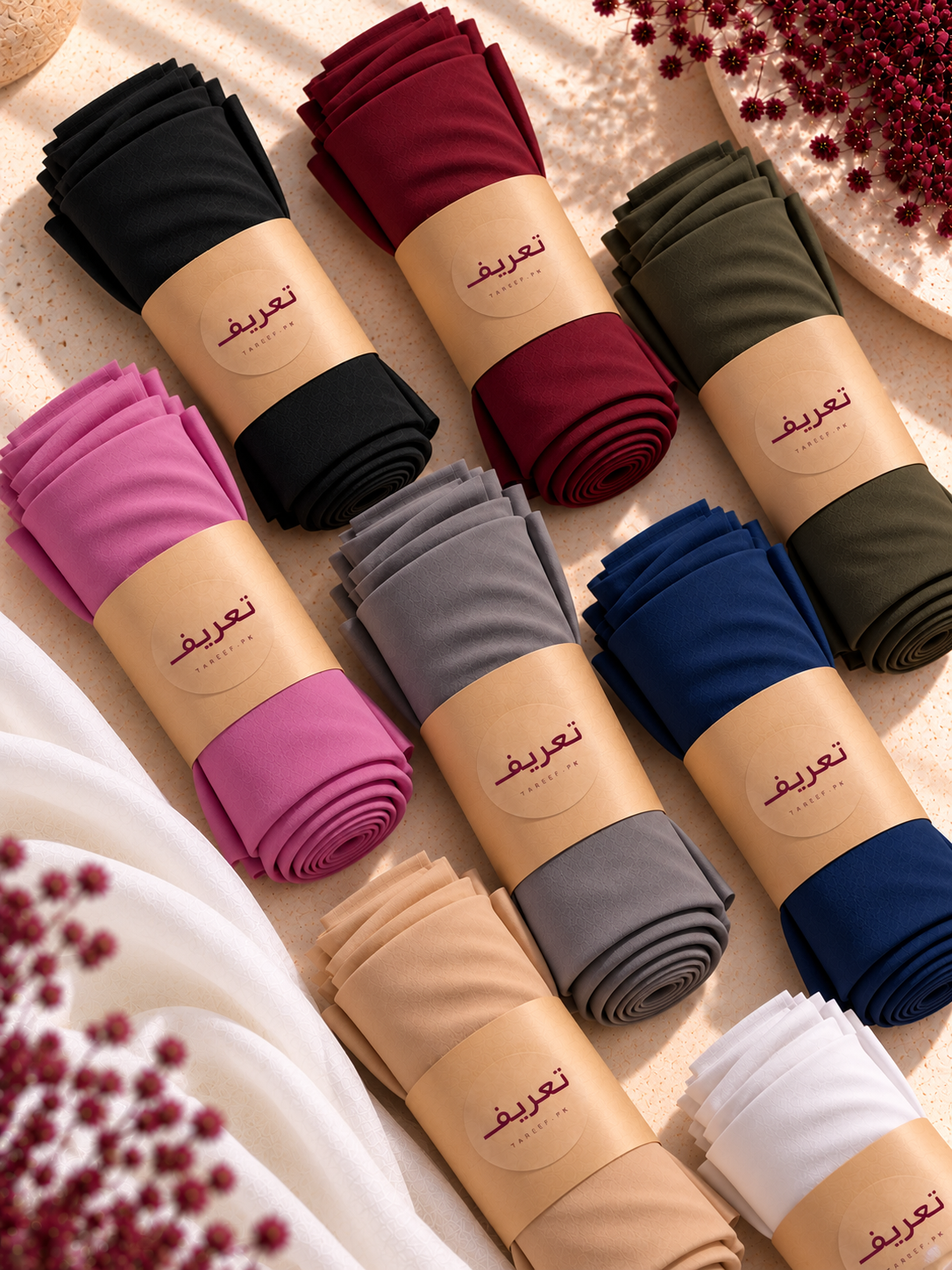 Multiple rolled hijabs in assorted colors, each wrapped with a tan paper band printed with the Arabic brand name 'TAREEF' arranged on a light beige textured surface with decorative floral props.